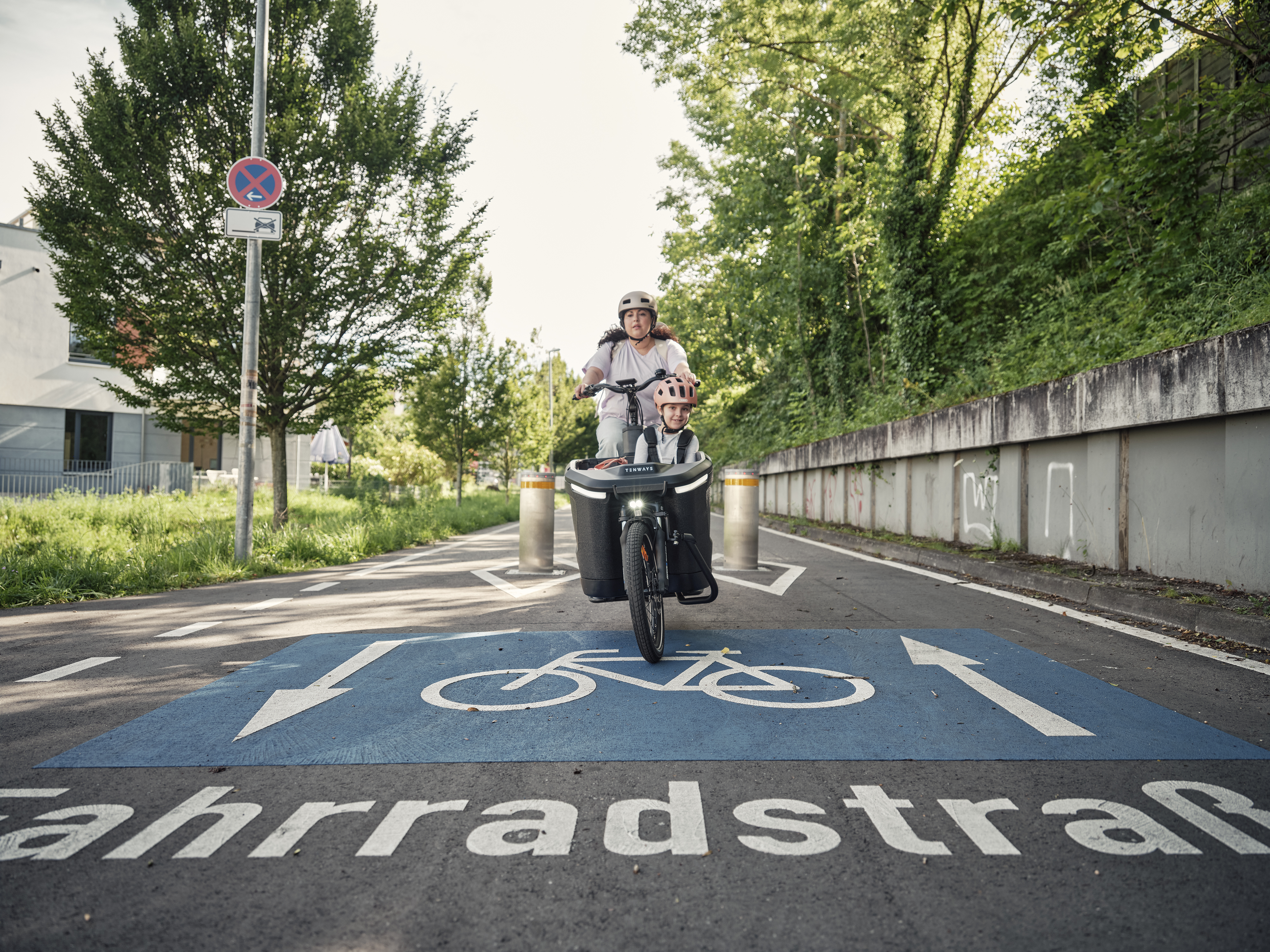 Mama mit Kind im Lastenrad fährt auf Fahrradstrasse zwischen Pollern hindurch